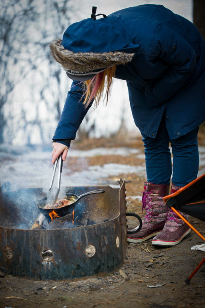 woman bends over, cooking in a skillet over a fire in Algonquin Provincial Park