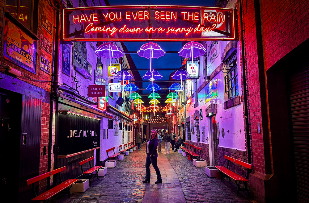 Belfast Cathedral Quarter Commercial Court in early evening. Author posing under sign that reads Have you ever seen the rain coming down on a sunny day?